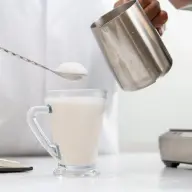 A glass half filled with sweet boba and ice, with milk tea being poured the rest of the way