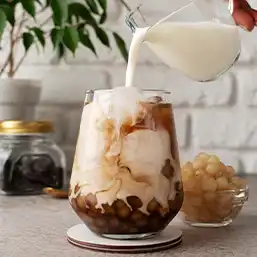 A glass half filled with sweet boba and ice, with milk tea being poured the rest of the way